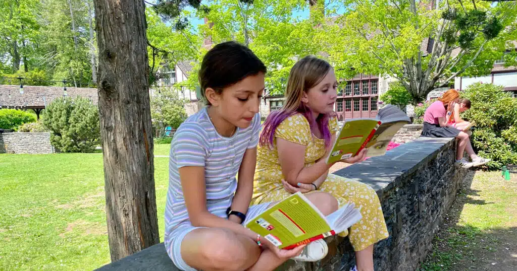 2 girls sitting on stone wall reading books
